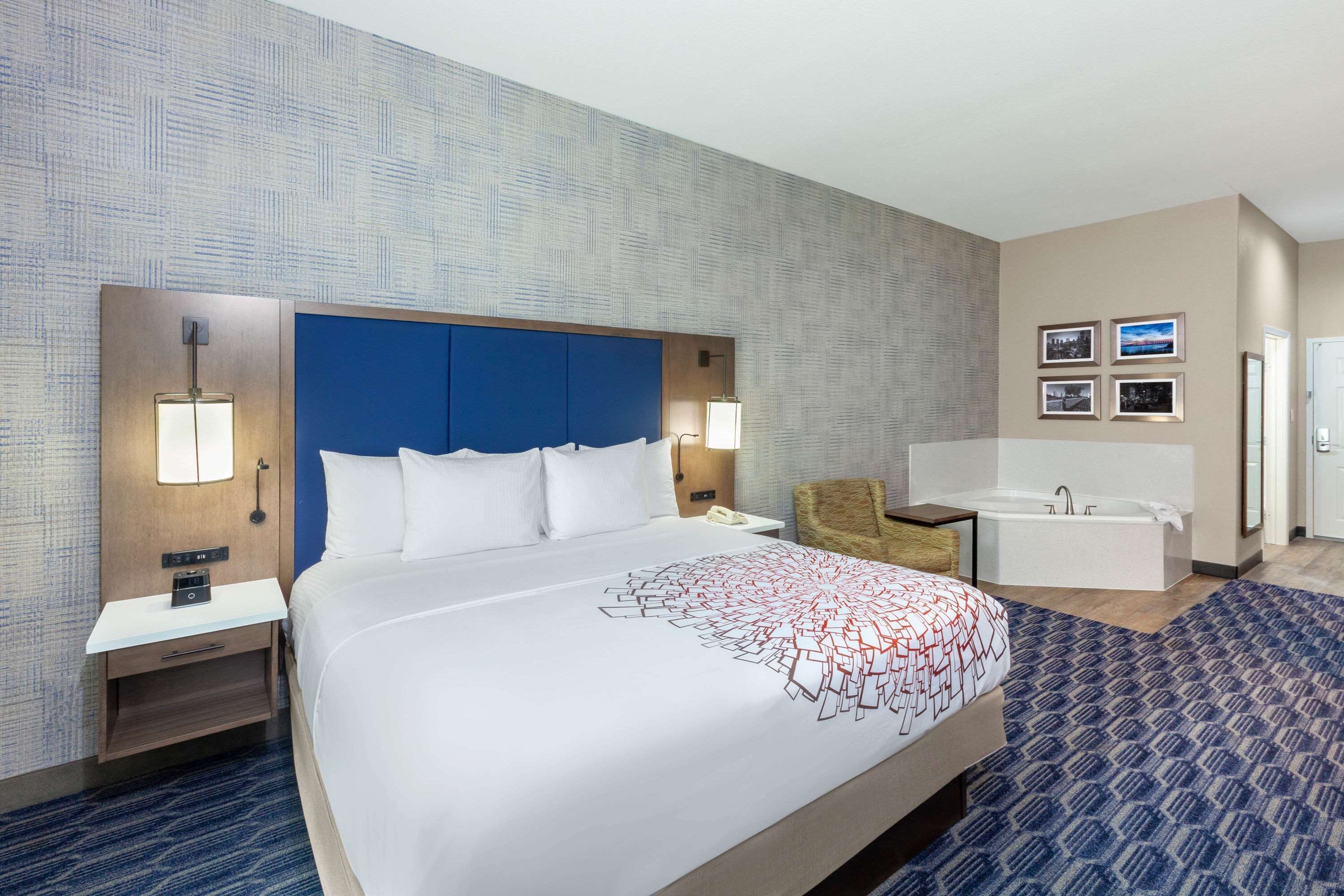 A hotel room with a large bed, a blue headboard, a sitting area with a chair, a small table, and a bathtub.