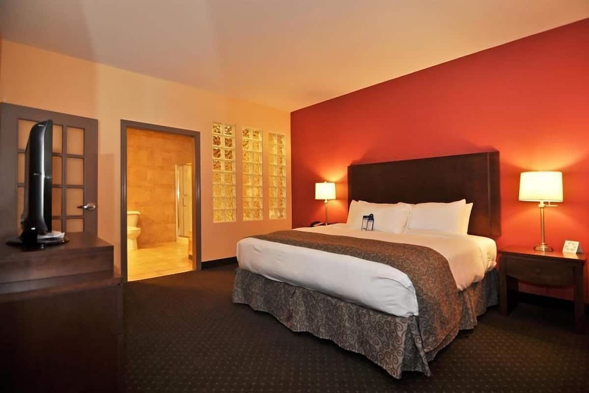 A hotel room with a large bed, two bedside lamps, a wooden headboard, a nightstand, and a door leading to a bathroom.