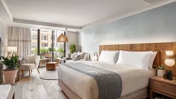 A modern hotel room with a large bed, a desk, a chair, a lamp, and a view of the city.