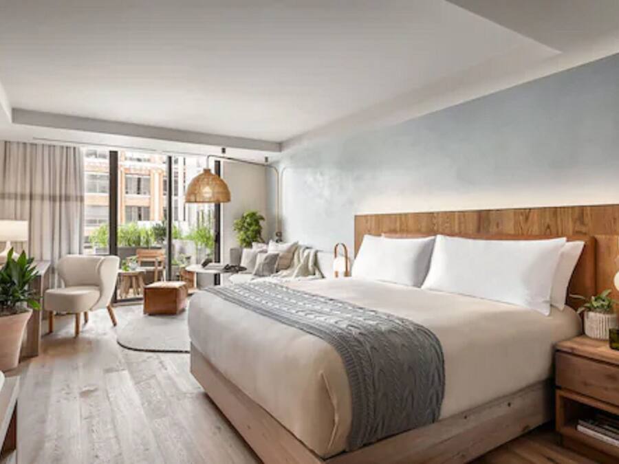 A modern hotel room with a large bed, a desk, a chair, a lamp, and a view of the city.