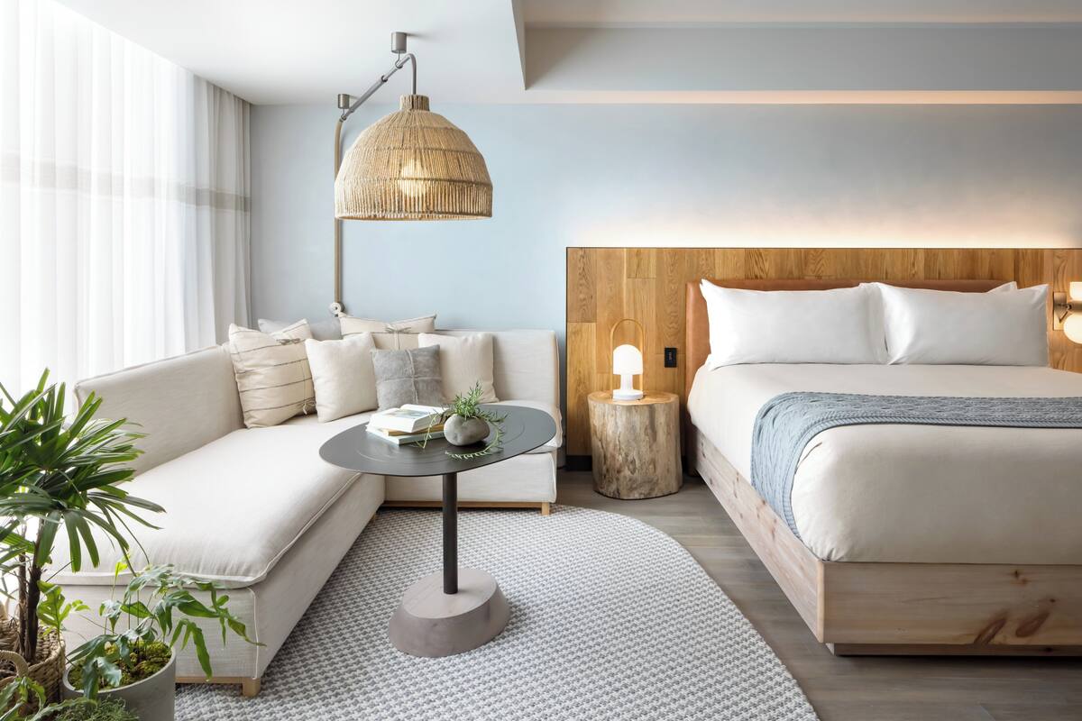 Une chambre d’hôtel moderne avec un canapé, une table basse ronde, un lit, une petite table d’appoint et une grande fenêtre.