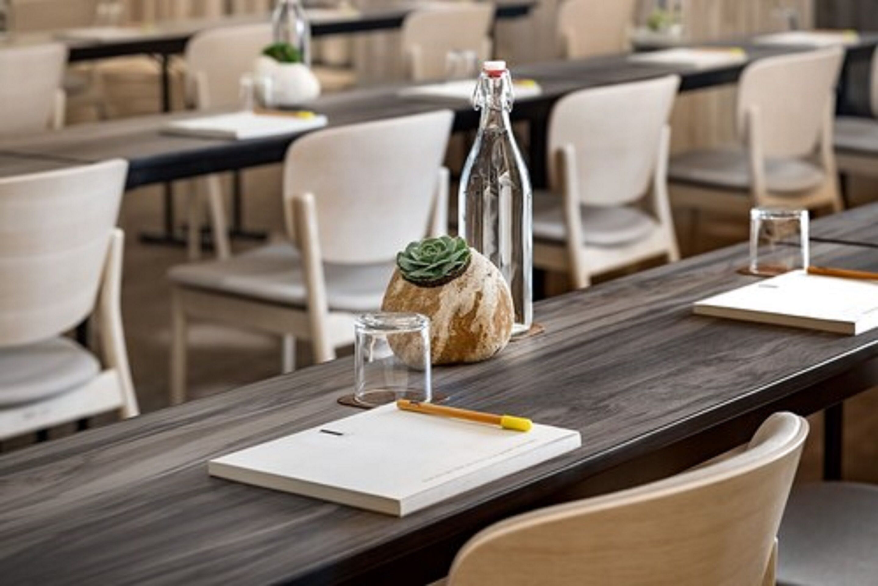 Une salle de conférence avec de longues tables, des chaises blanches, une bouteille en verre, un petit verre et un carnet.