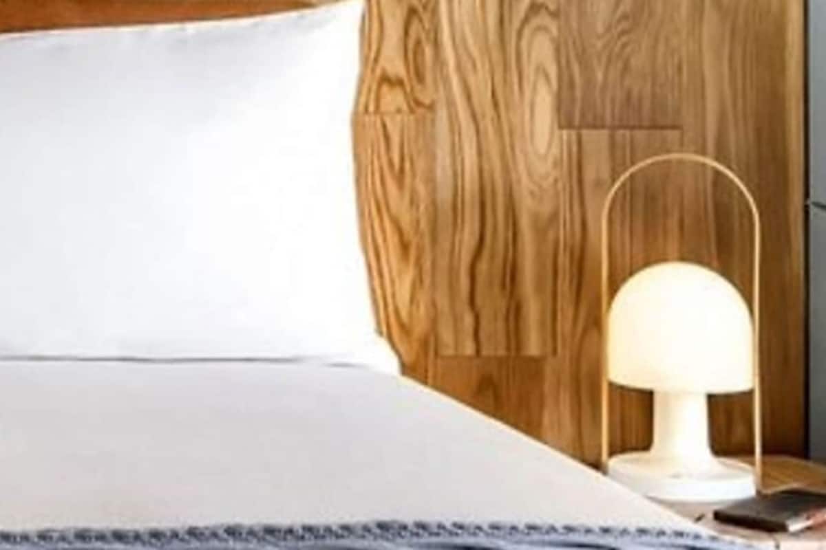 A neatly made bed with a white pillow and a wooden headboard. A small table with a lamp and a book is beside the bed.