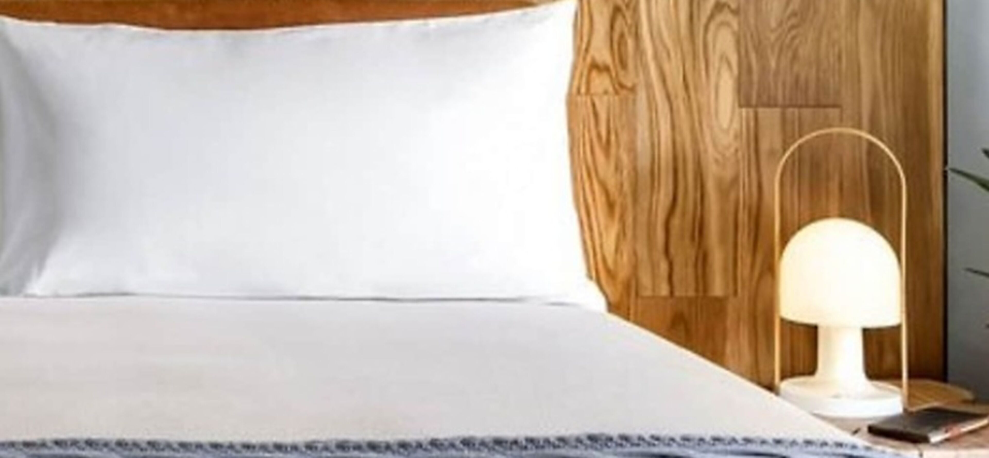 A bed with a white pillow, a wooden headboard, a bedside lamp, and a smartphone on a bedside table.