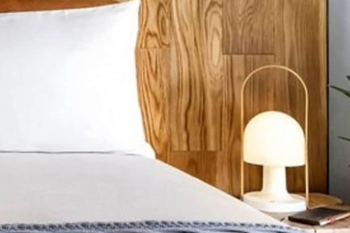 Un lit bien fait, avec un oreiller blanc et une tête de lit en bois. Une petite table avec une lampe et un livre se trouve à côté du lit.
