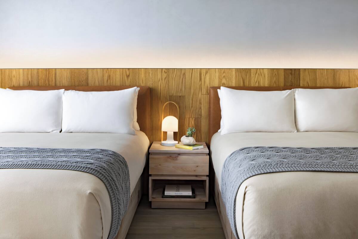 Une chambre d’hôtel avec deux lits, des têtes de lit en bois, du linge de lit blanc, une table de chevet avec une lampe et une petite plante.