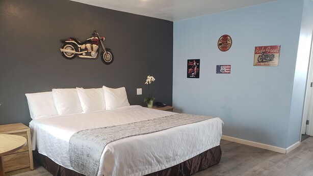 A neatly made bed with a white comforter and brown skirt, a wooden nightstand with a lamp, and a wall with vintage motorcycle and Route 66 memorabilia. - Deluxe Inn Motel (Seligman)