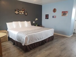 A neatly made bed with a white comforter and brown skirt, a wooden nightstand with a lamp, and a wall with vintage motorcycle and Route 66 memorabilia.