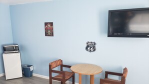 A small room with a round wooden table and two chairs, a microwave, a trash bin, and a television mounted on the wall. - Deluxe Inn Motel (Seligman)