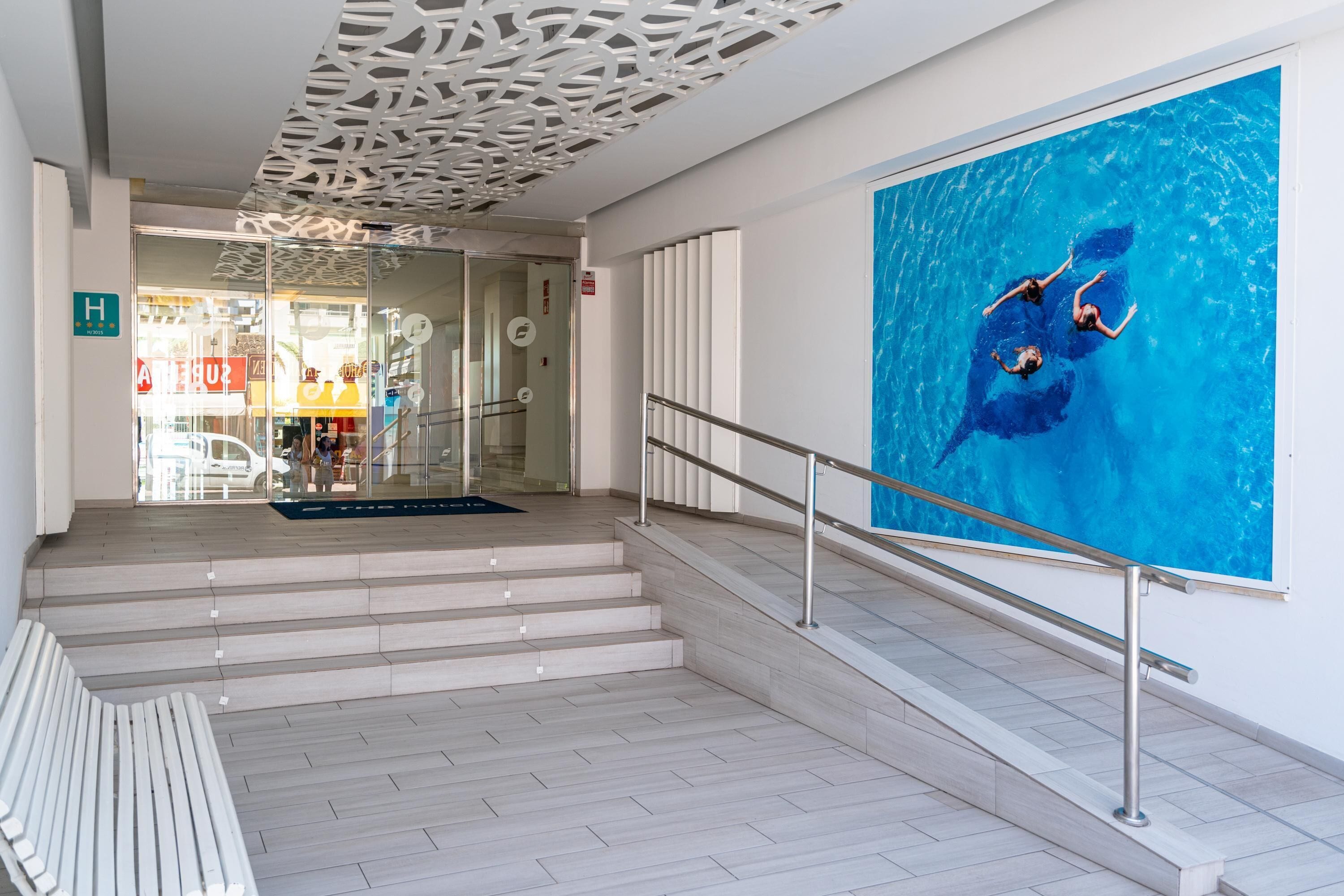 Un vestíbulo de hotel con una escalera, una entrada por una puerta de cristal y un gran mural azul de nadadores.
