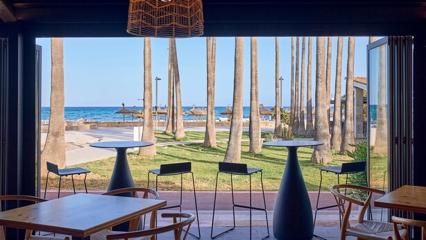 An indoor seating area with tables and chairs, overlooking a beach with palm trees and umbrellas. - Hotel THB Sa Coma Platja (Sant Llorenc des Cardassar)