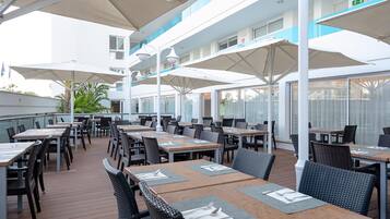 Un área de comedor al aire libre con varias mesas y sillas, cubierta por sombrillas, y vista al exterior del edificio.