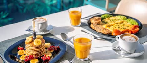 A breakfast setting with pancakes, fruit, juice, and coffee on a table by a pool.