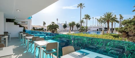 A rooftop dining area with tables and chairs, overlooking a pool and palm trees.