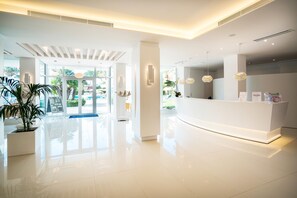 A modern hotel lobby with a reception desk, large windows, and a potted plant.