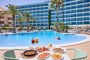Zona de comedor junto a la piscina con vistas a un edificio de hotel, palmeras y personas nadando.