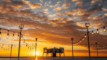 Vista del atardecer con una mesa y sillas sobre un muelle, luces colgantes y un farol al fondo.