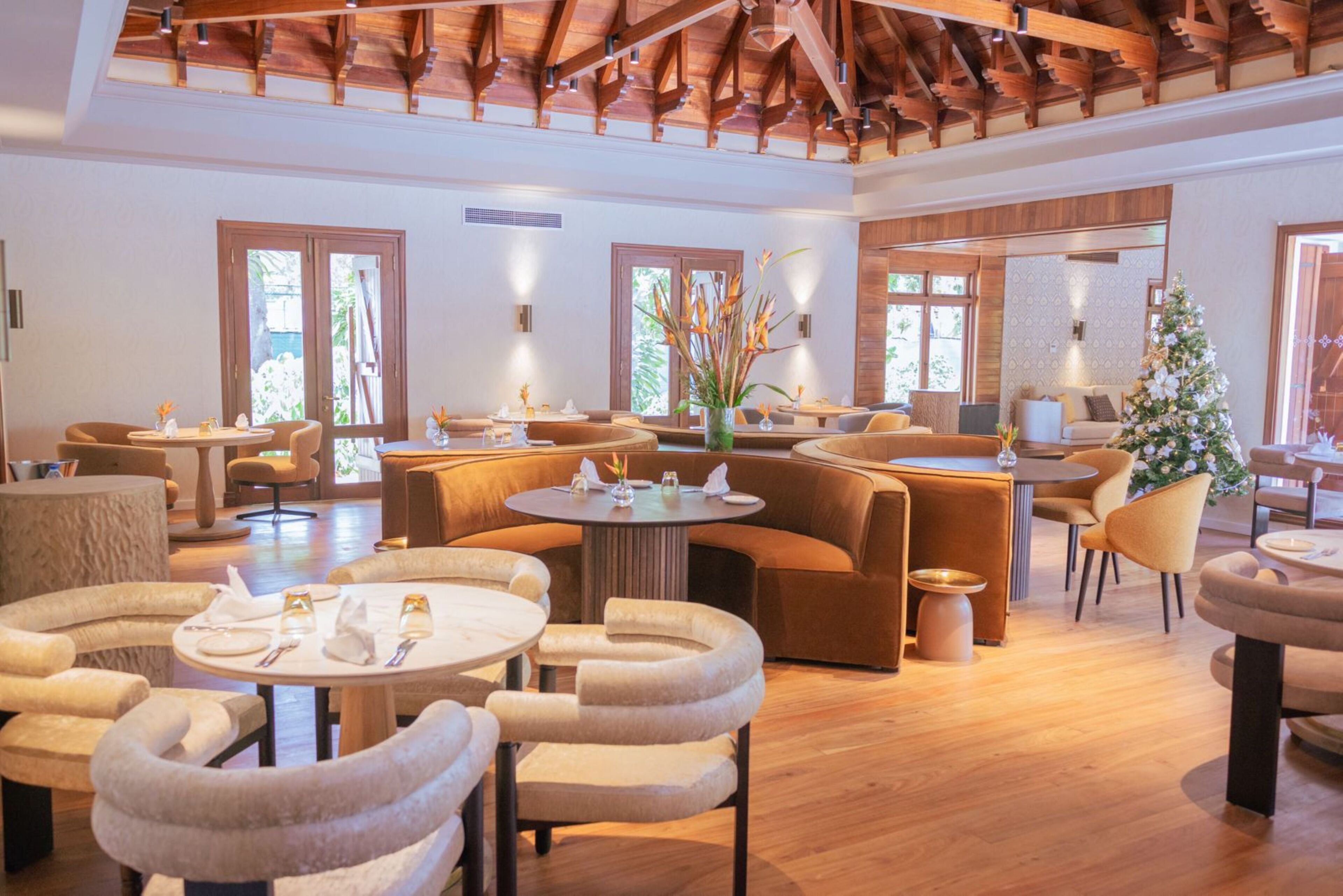 A spacious, well-lit lounge area with multiple seating options including sofas and round tables, wooden flooring, and large windows allowing natural light.
