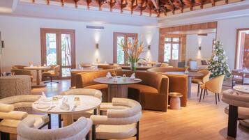 A spacious, well-lit lounge area with multiple seating options including sofas and round tables, wooden flooring, and large windows allowing natural light.