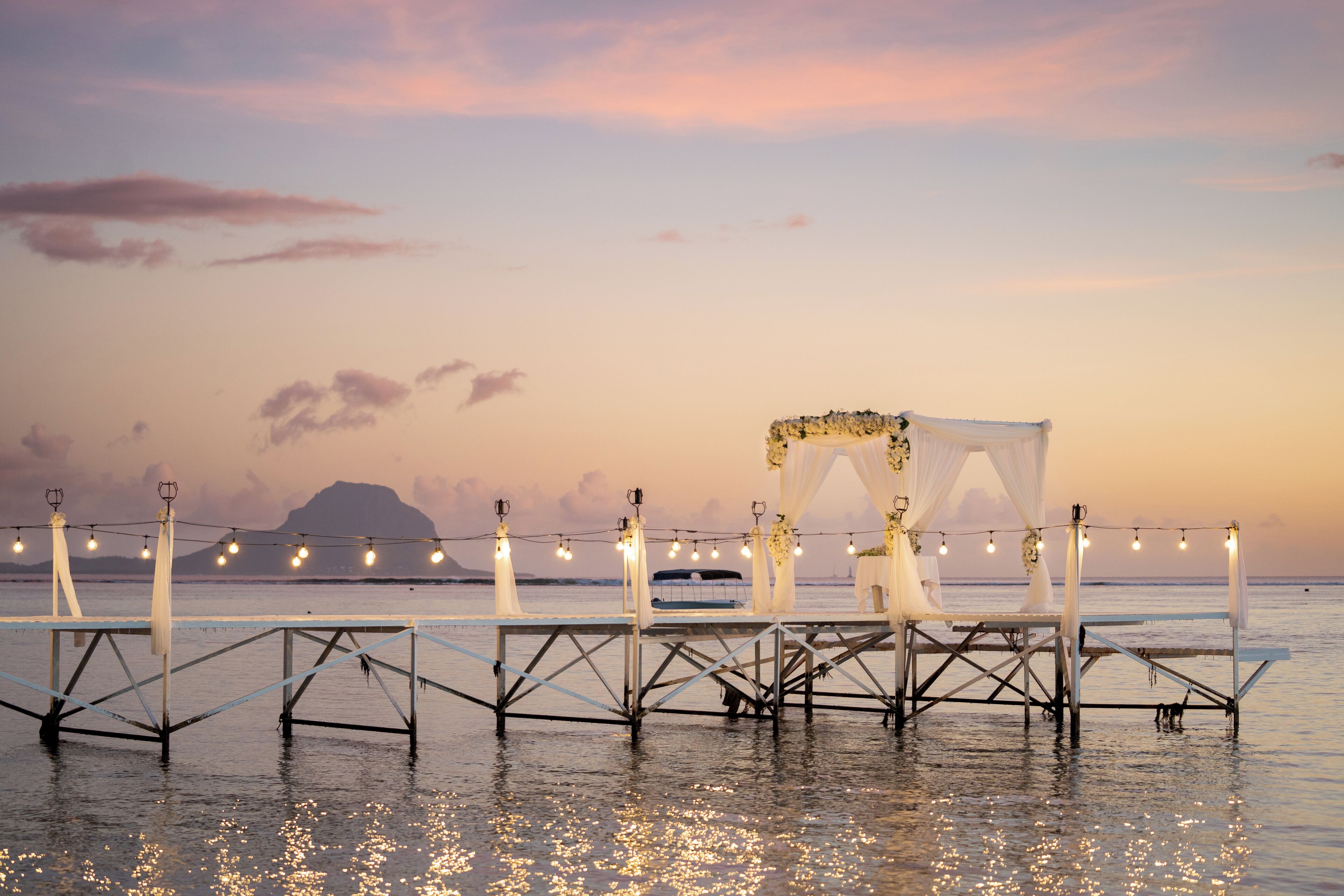 A pier with a canopy and string lights, overlooking a calm body of water with a sunset in the background.