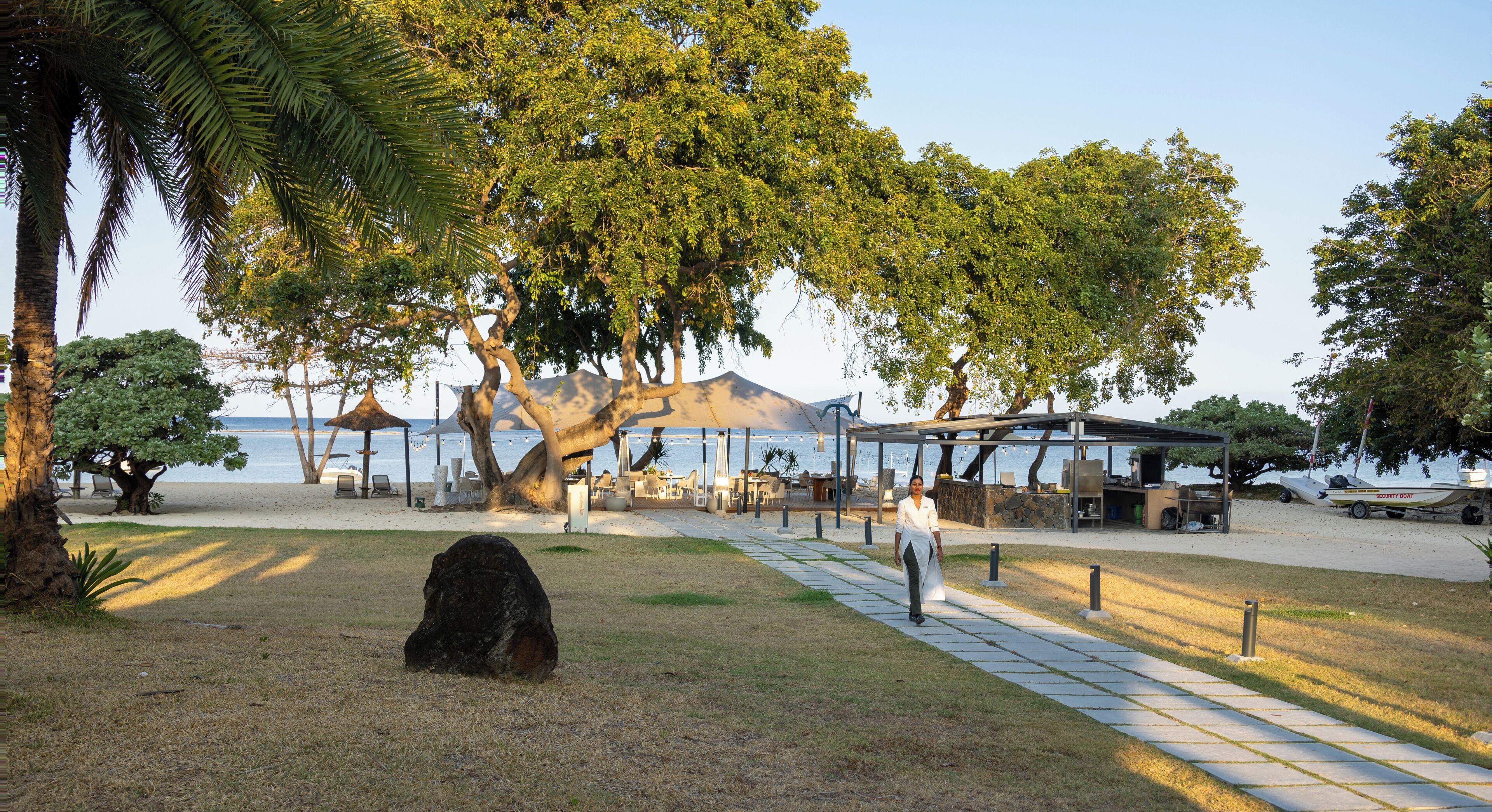 Ein Park direkt am Strand mit einem großen Baum, einer überdachten Sitzgelegenheit und Blick aufs Meer.