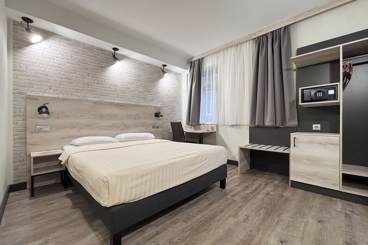 A modern hotel room with a large bed, a desk, a chair, a wardrobe, and a small table.