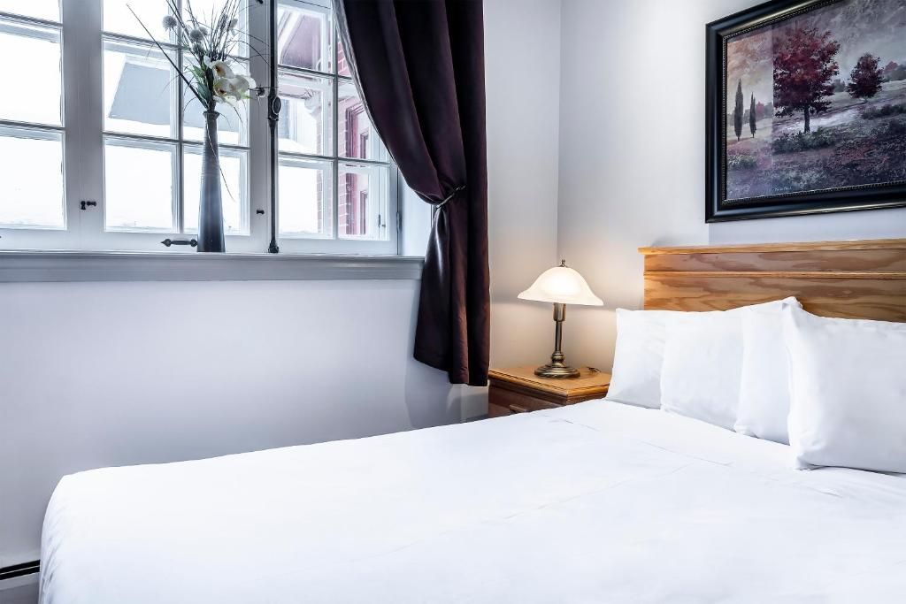 Quarto de hotel com uma cama, mesa de cabeceira com abajur, janela com cortinas e um quadro na parede.