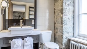 A bathroom with a stone wall, a white toilet, a sink with a mirror, and a window. - Hotel Louisbourg (Québec City)