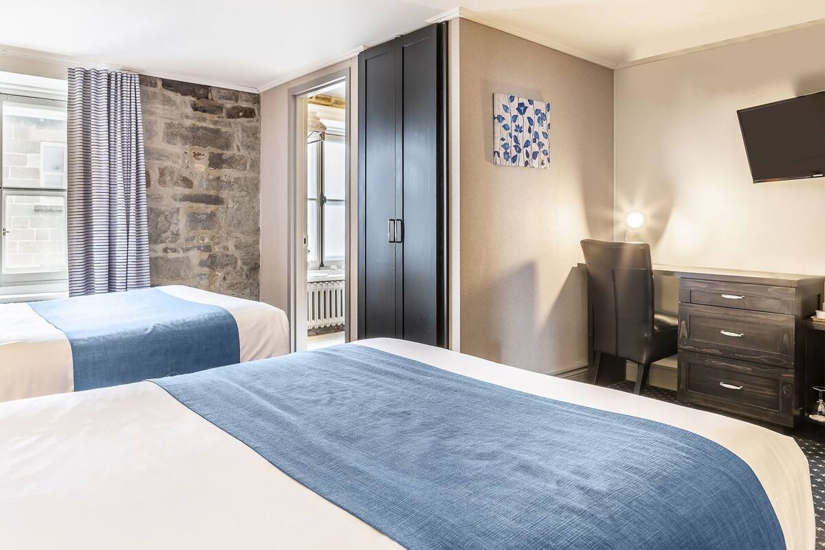 Une chambre d’hôtel avec deux lits, un mur en pierre, une armoire sombre, un bureau et un téléviseur à écran plat.