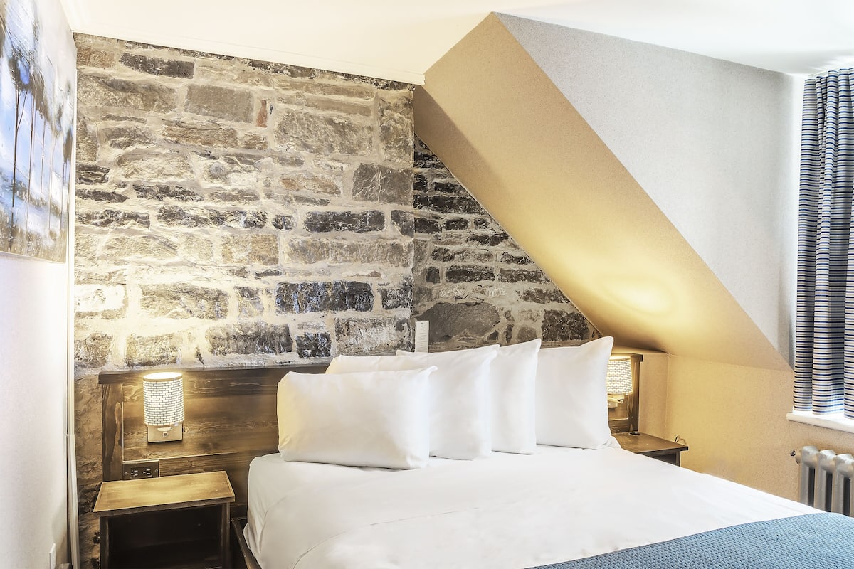 Une chambre avec un mur en pierre, un plafond en pente, un lit avec une literie blanche et une table de chevet avec une lampe.
