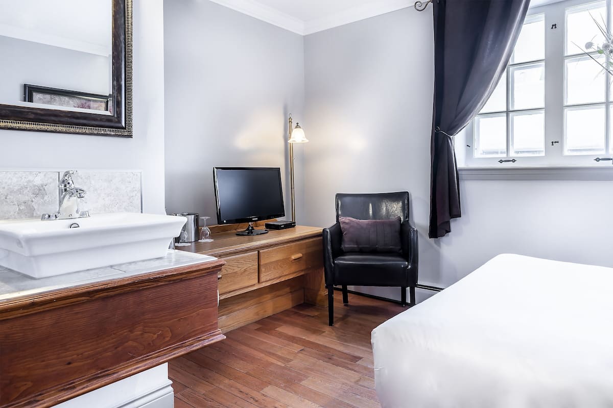 Une chambre d’hôtel avec un bureau en bois, une chaise noire, un lit, un lavabo blanc et une grande fenêtre avec des rideaux.