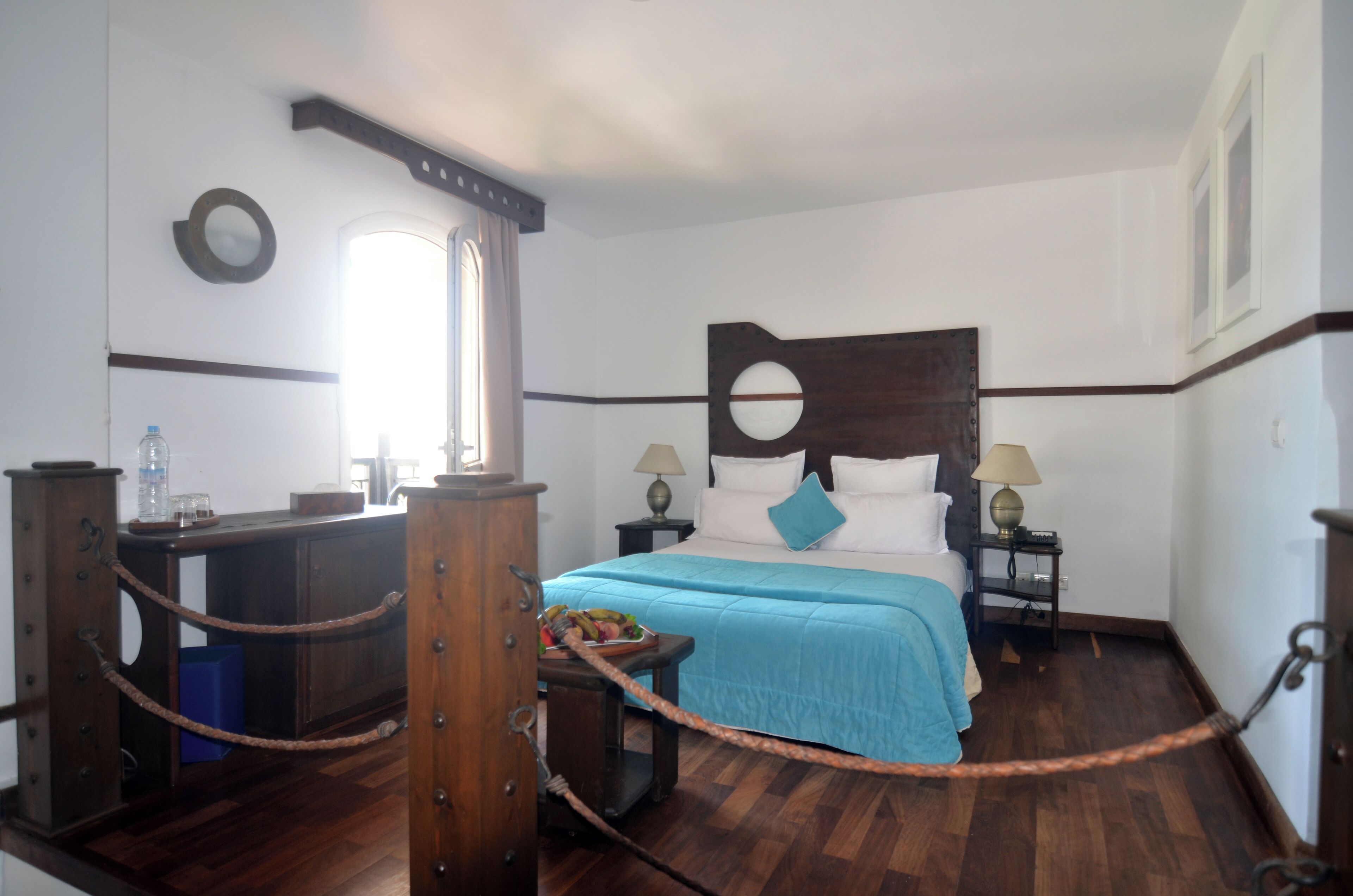 Habitación de hotel con cabecera de madera, cama con colcha azul, dos mesitas de noche con lámparas, un escritorio de madera con silla y una ventana con cortinas.
