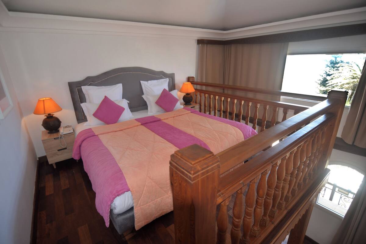 A hotel room with a wooden bed frame, a pink and peach quilt, two bedside lamps, and a view of the outdoors through a window.