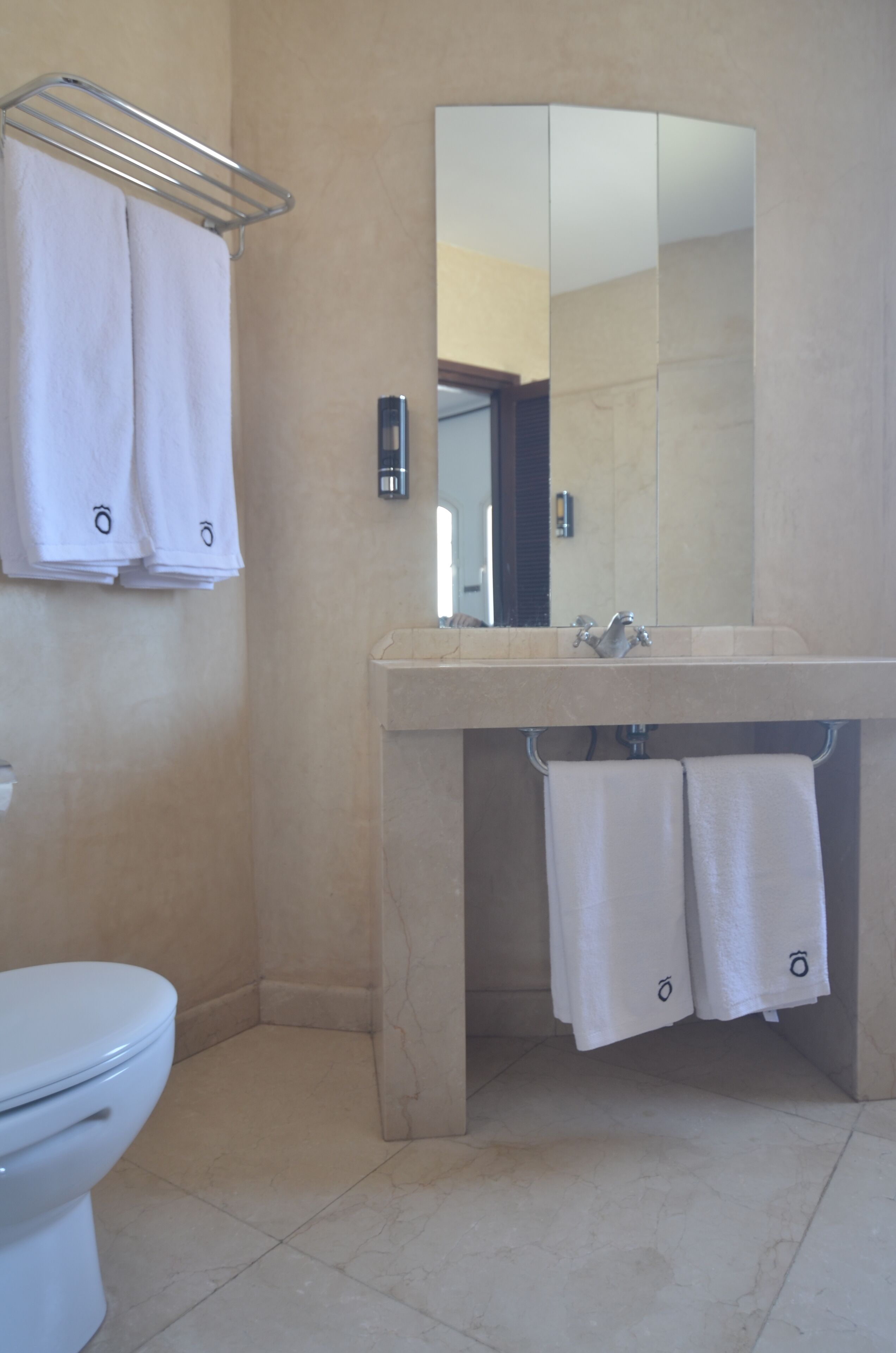 Un baño con inodoro, lavamanos con espejo y dos toallas colgadas en un perchero.