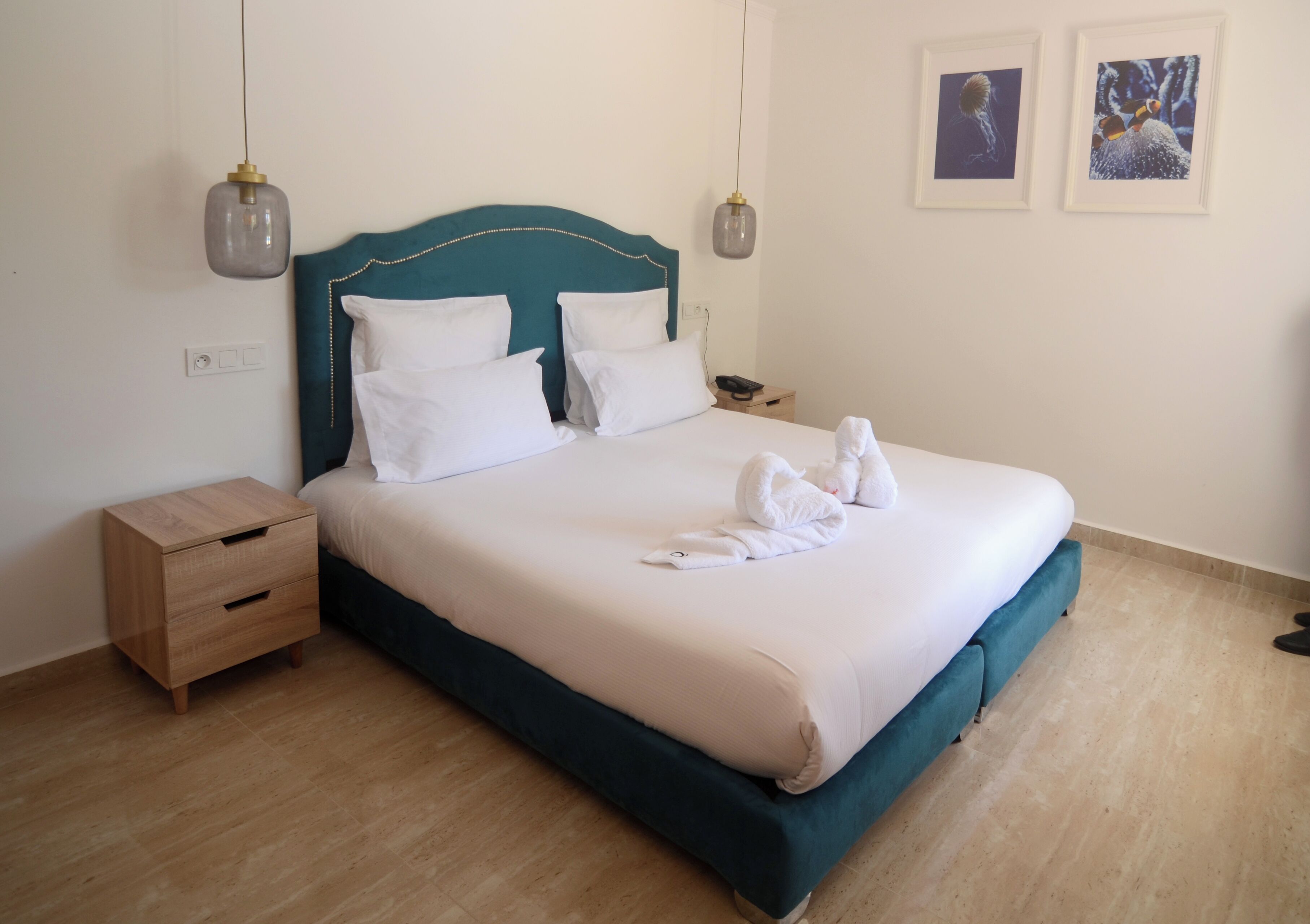 A neatly made bed with white linens and a teal headboard, a wooden nightstand with drawers, and two framed pictures on the wall.