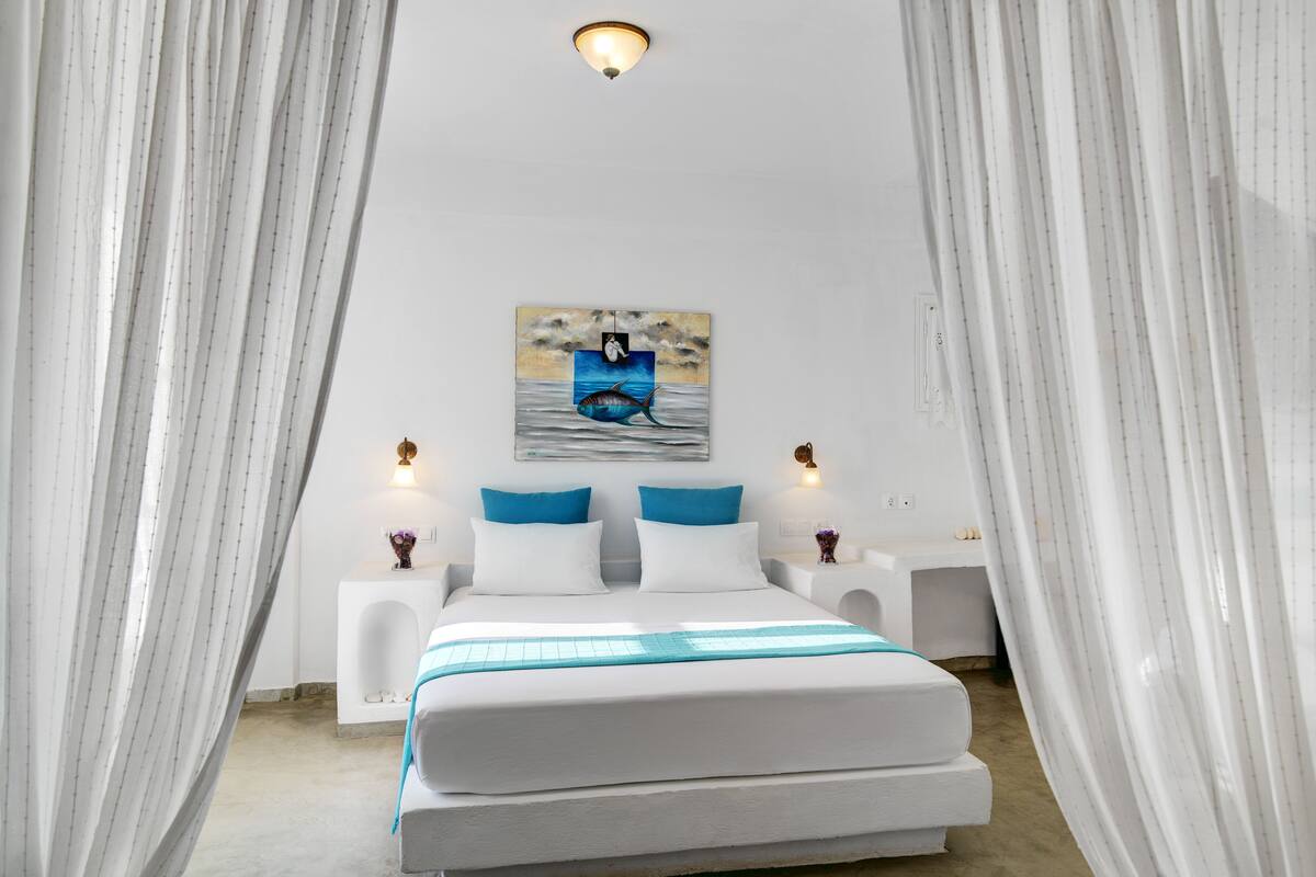 A bedroom with a bed, white curtains, a painting of a fish, and two bedside lamps.
