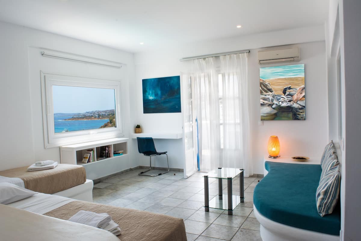 A modern hotel room with a large window offering a view of the sea, a cozy seating area with a sofa and a chair, and a small table with a lamp.