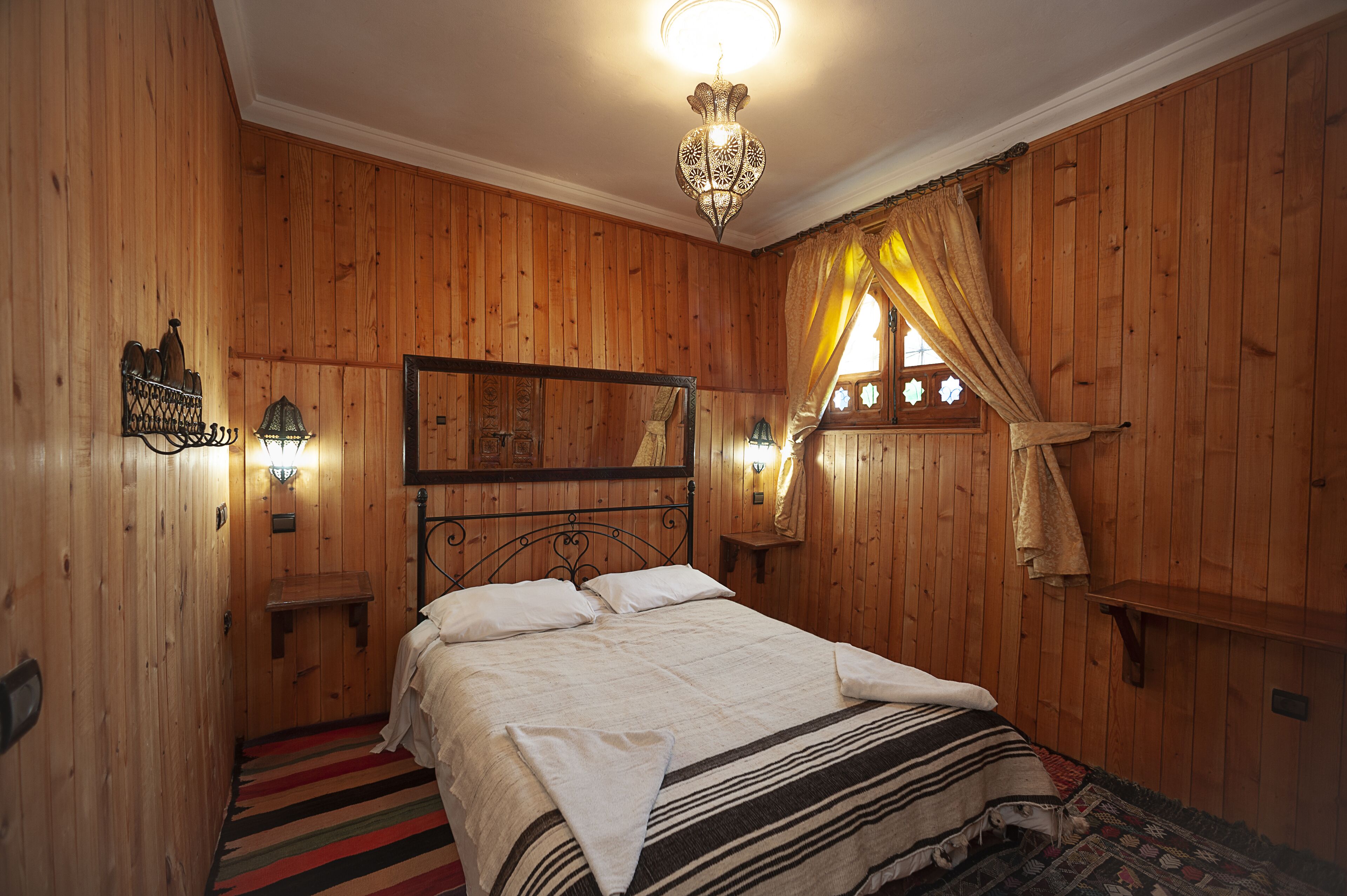 Una habitación con paneles de madera, una cama, un colgante de luz y una ventana con cortinas.