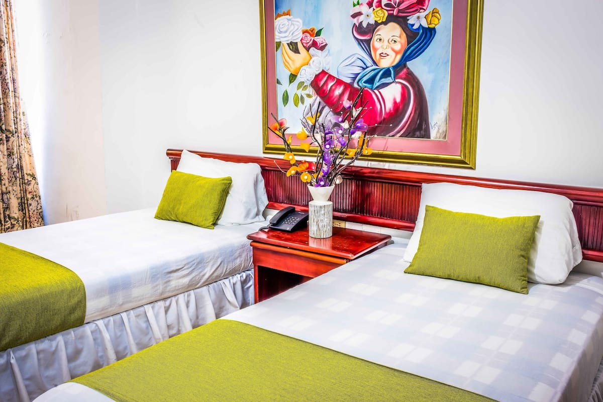 Ein Hotelzimmer mit zwei Betten, beide mit grüner Bettwäsche und weißen Kissen. Darüber an der Wand hängt ein Gemälde, auf dem Nachttisch steht eine Vase mit Blumen und ein Telefon.