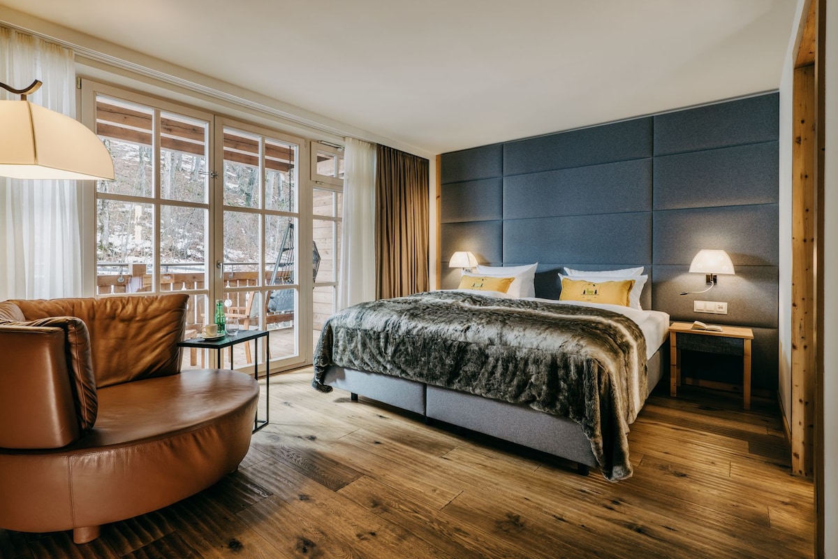 A modern bedroom with a large bed, a leather armchair, and a view of the outdoors through large windows.