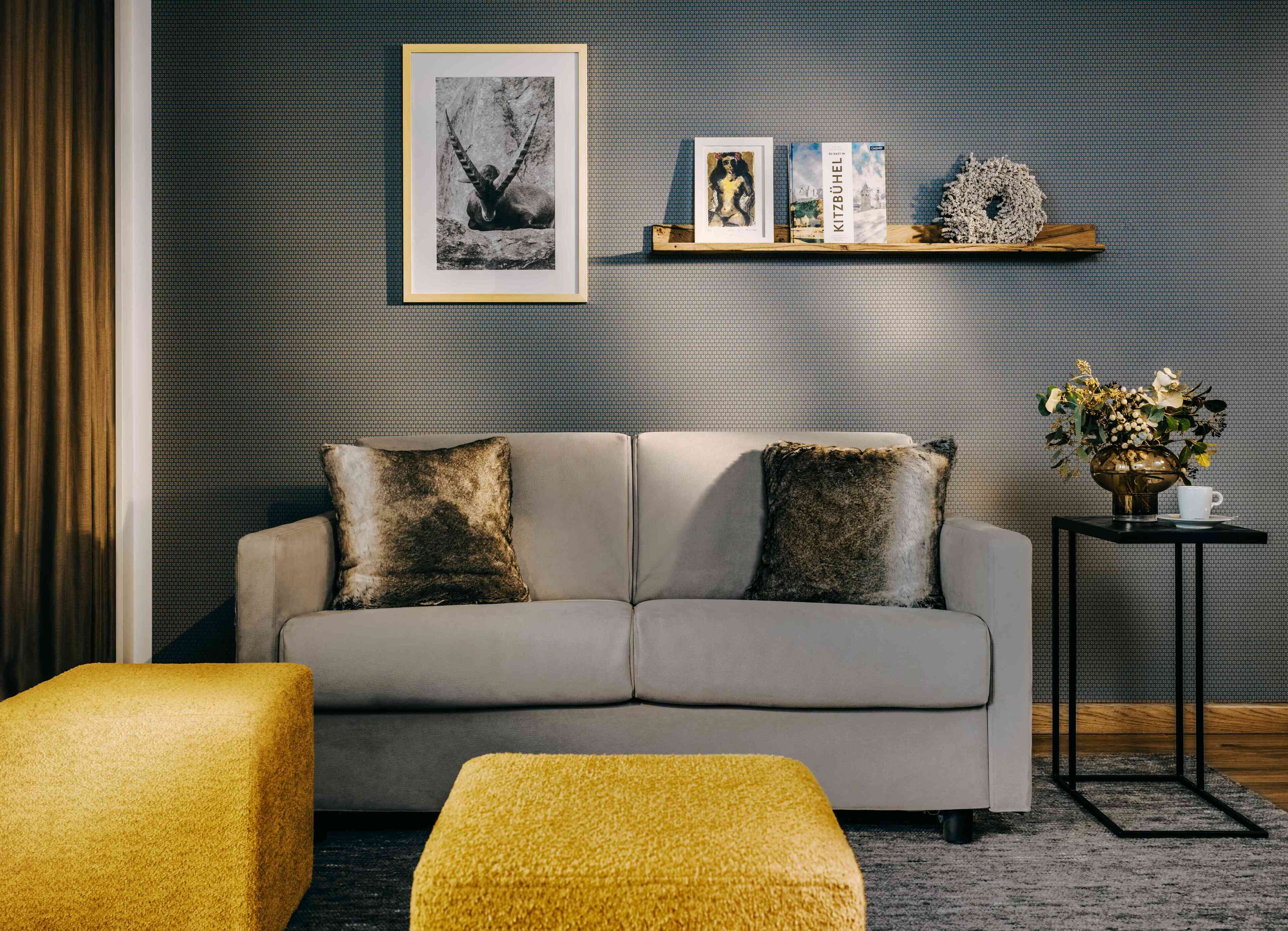 A modern living room with a grey sofa, two decorative pillows, a yellow ottoman, a framed picture, a book, and a small table with a vase and cup.