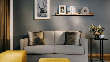 A modern living room with a grey sofa, two decorative pillows, a yellow ottoman, a framed picture, a book, and a small table with a vase and cup.