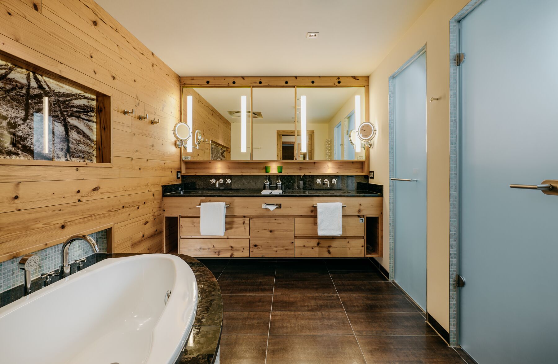 A modern bathroom with wooden paneling, a large bathtub, a double vanity with mirrors, and a blue door.