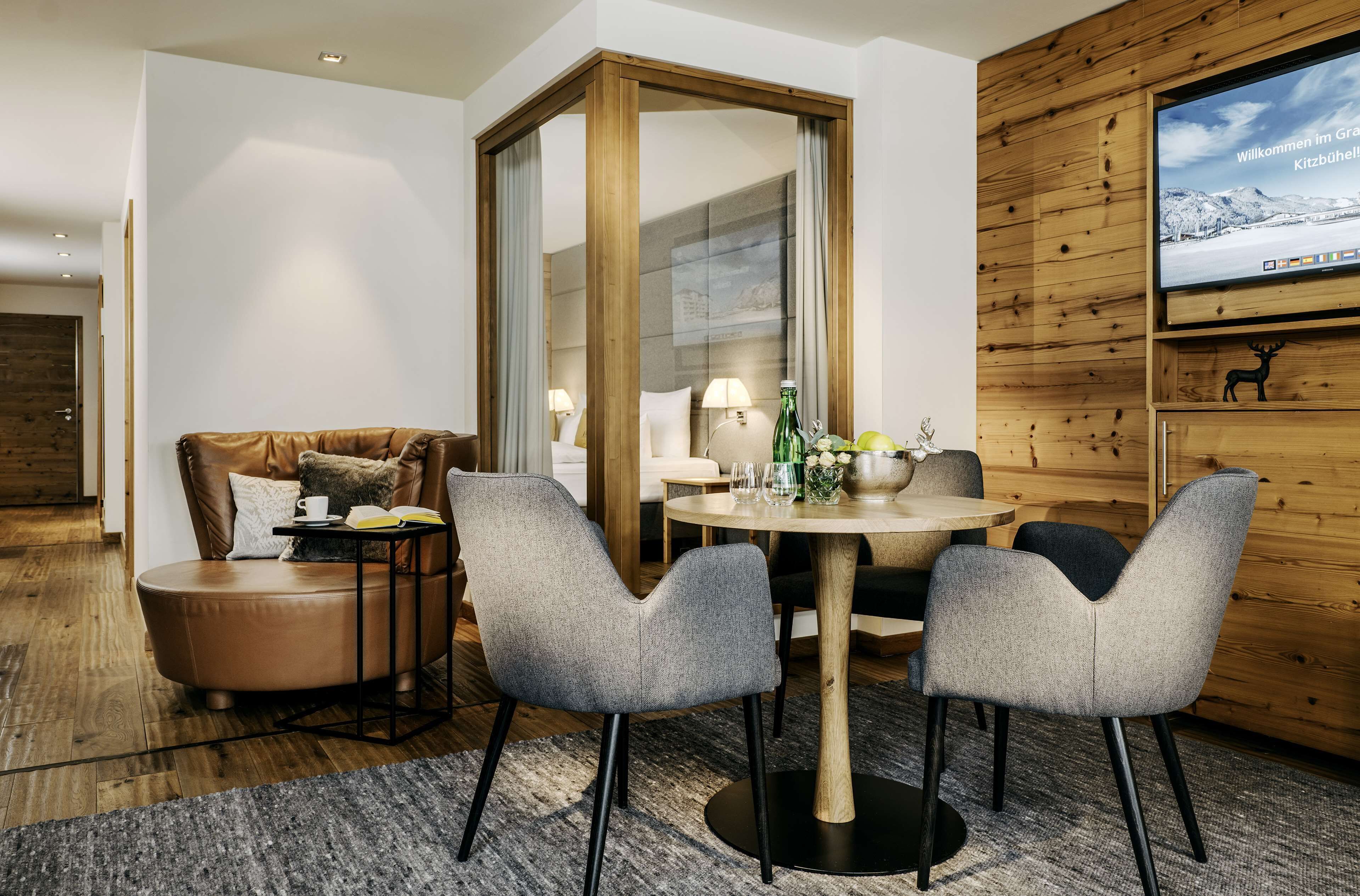 Une chambre d’hôtel moderne avec un mur d’accent en bois, un grand téléviseur à écran plat, un canapé en cuir, une table de salle à manger ronde avec des chaises et une cloison en verre.