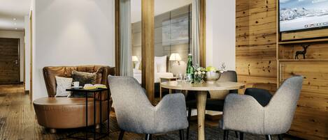 Une chambre d’hôtel moderne avec un mur d’accent en bois, un grand téléviseur à écran plat, un canapé en cuir, une table de salle à manger ronde avec des chaises et une cloison en verre.