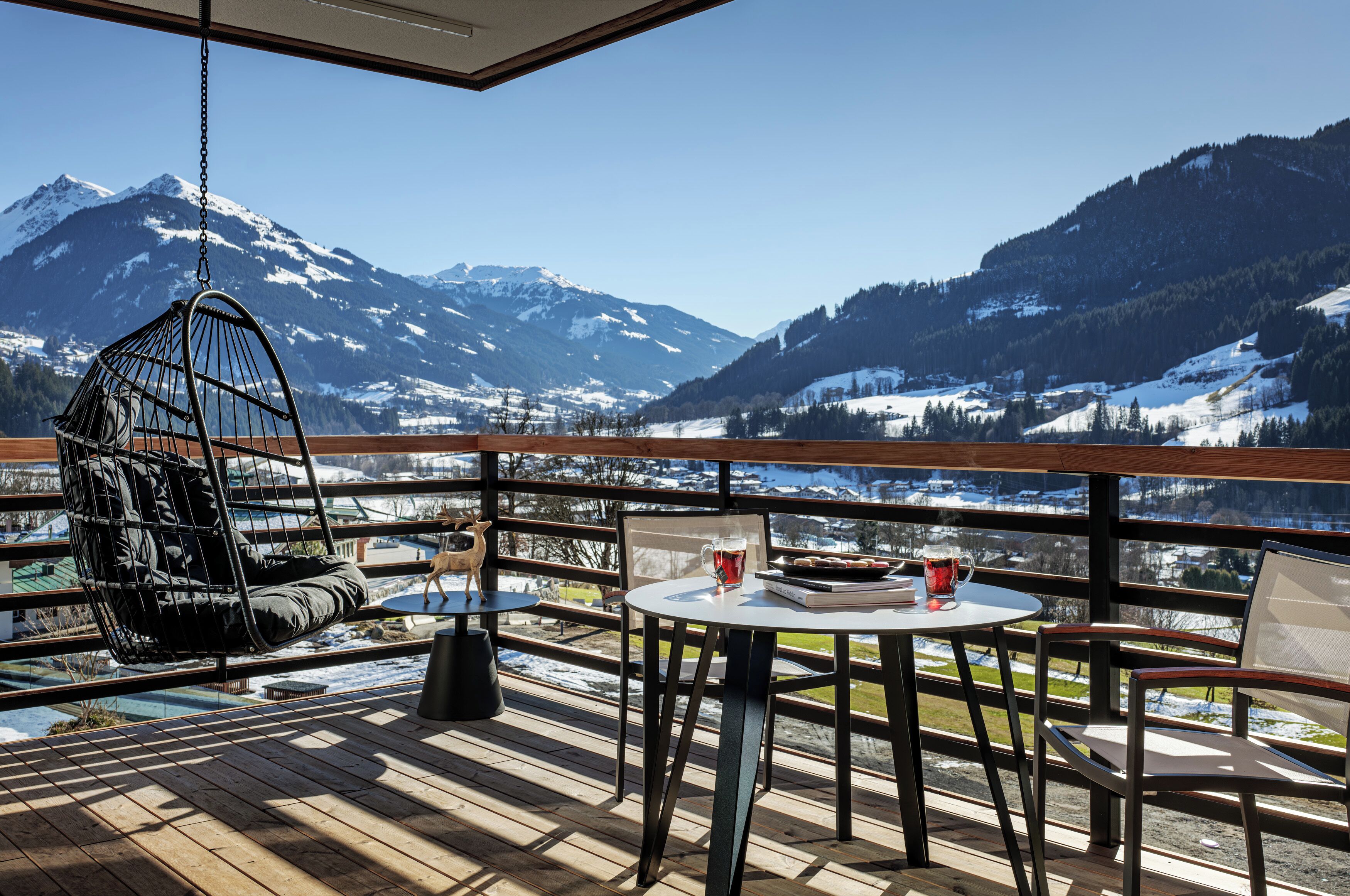 A wooden deck with a hanging chair, a table with a book and a mug, and a view of snow-covered mountains.