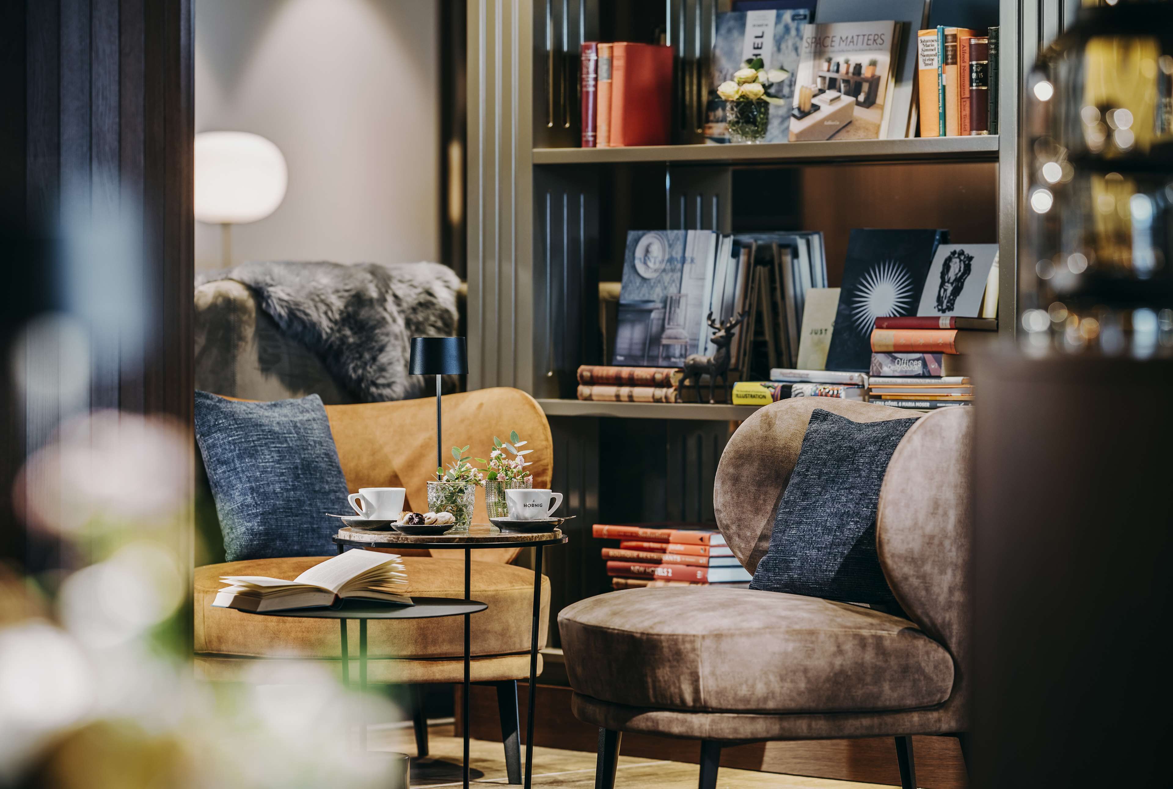 Een gezellig hoekje om in te lezen, met een comfortabele fauteuil, een klein bijzettafeltje met een boek en een kop, en een boekenplank vol boeken en decoratieve objecten.