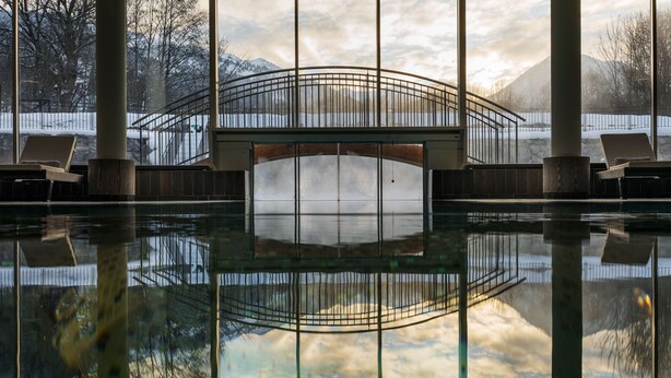 Una piscina interior tranquila con un puente de cristal, sillas de descanso y vistas nevadas al exterior.