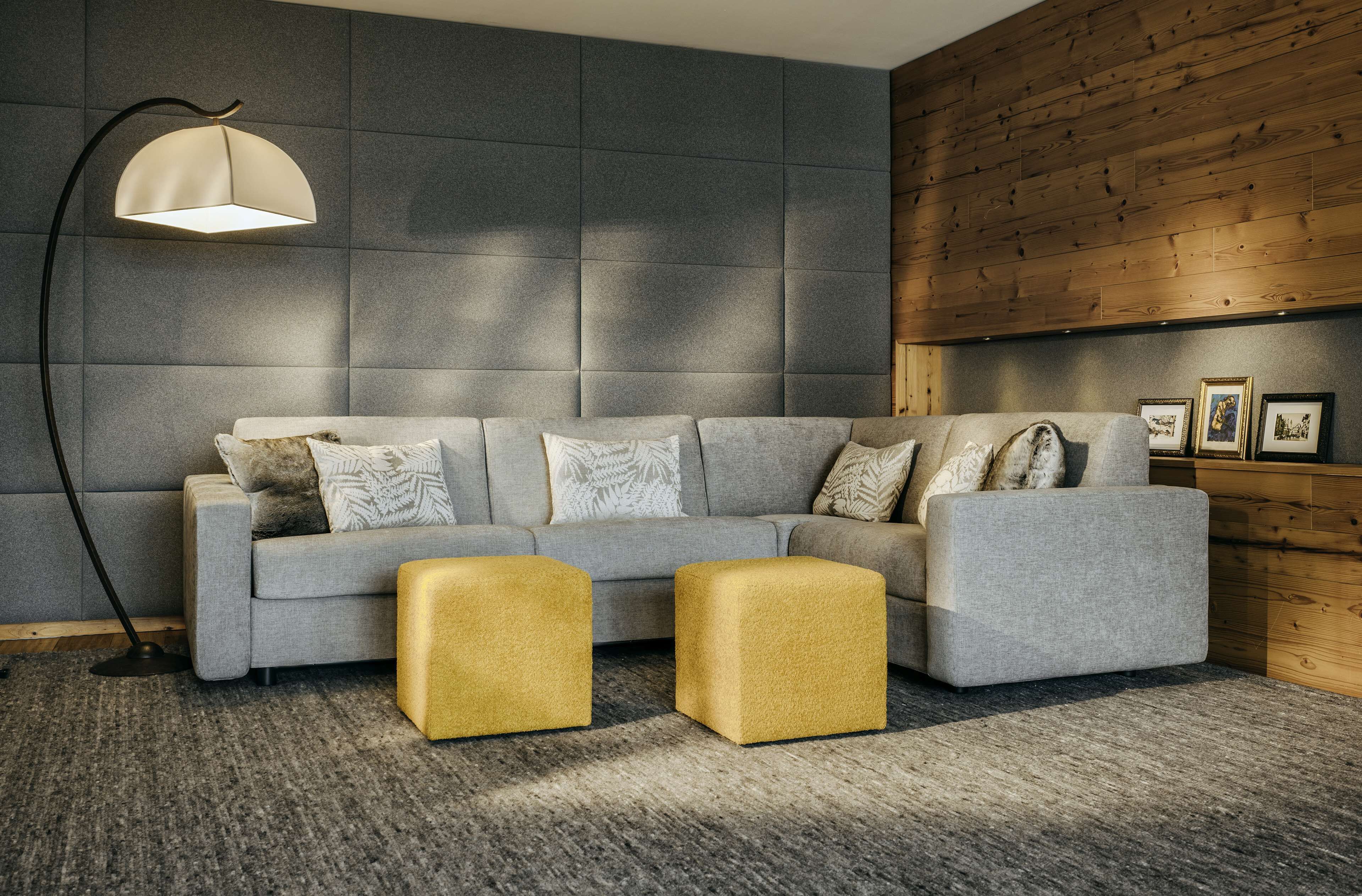 Een moderne woonkamer met een grijze bank, twee gele poefjes, een vloerlamp en een houten wandpaneel.