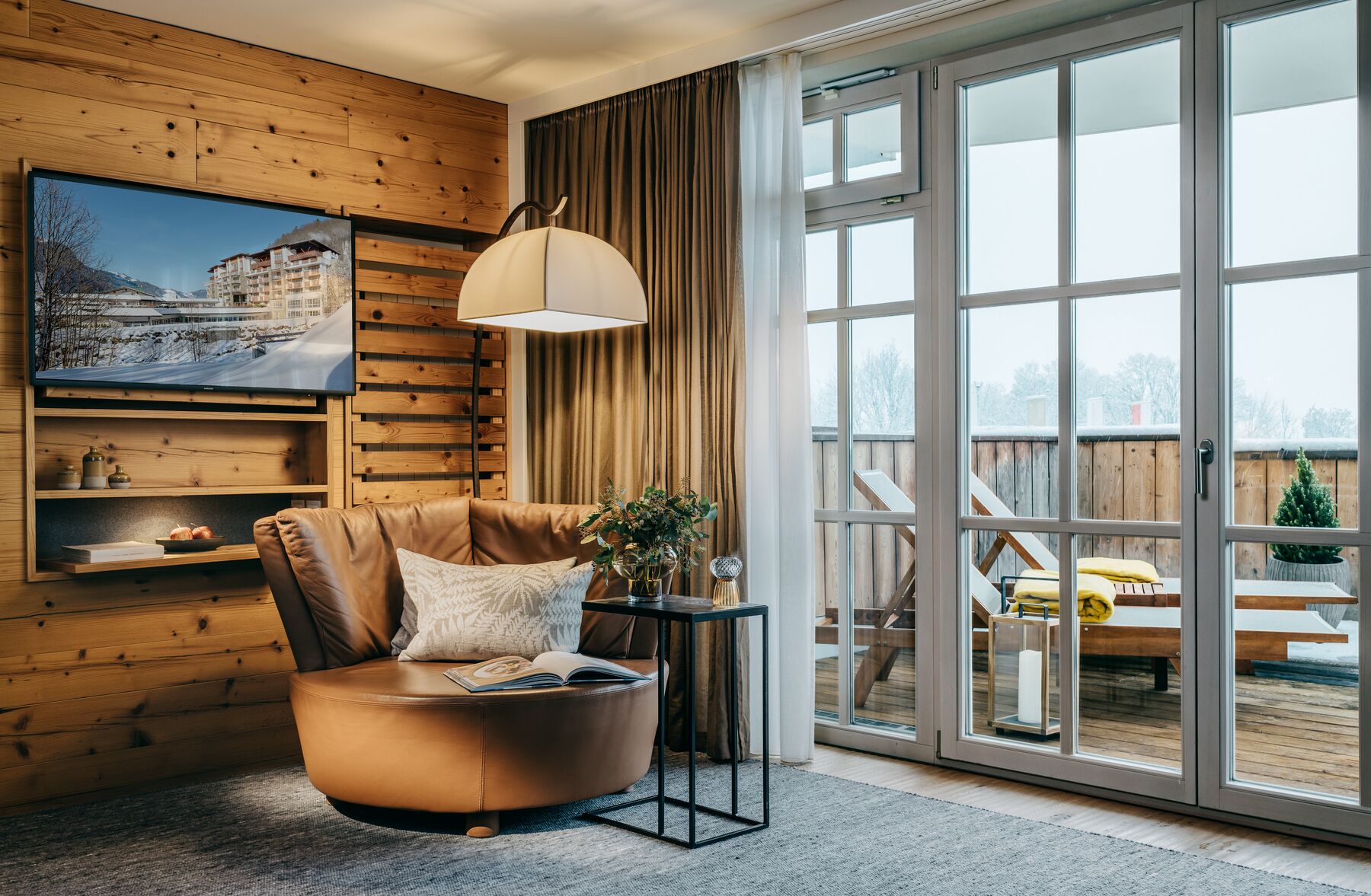 Un salon chaleureux avec un fauteuil en cuir, une petite table d’appoint ornée d’une plante et une grande fenêtre donnant sur une terrasse et sur du mobilier d’extérieur.